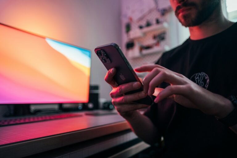 A hand holding a smartphone in front of a desk monitor
