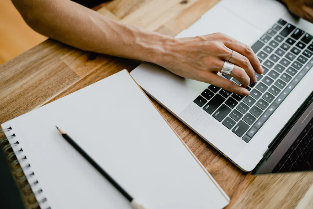 Hands typing on a laptop beside a notebook